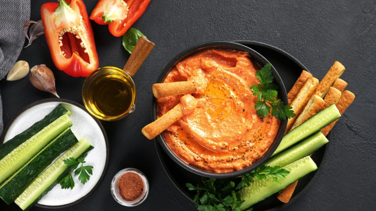 Greek feta spread Htipiti. Roasted red pepper, olive oil and feta dip served with sliced veggies and breadsticks, top view, dark background