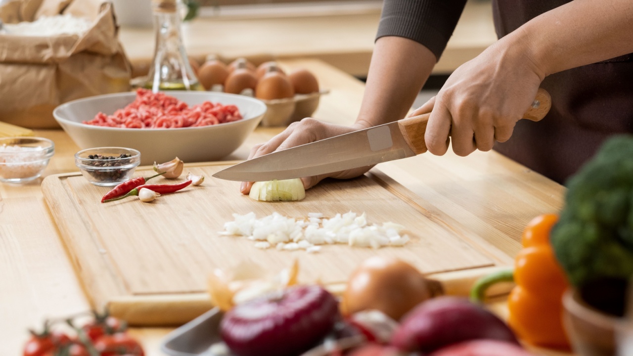 Hands of young female chopping fresh onion on wooden board while preparing italian pasta with vegetables and minced meat