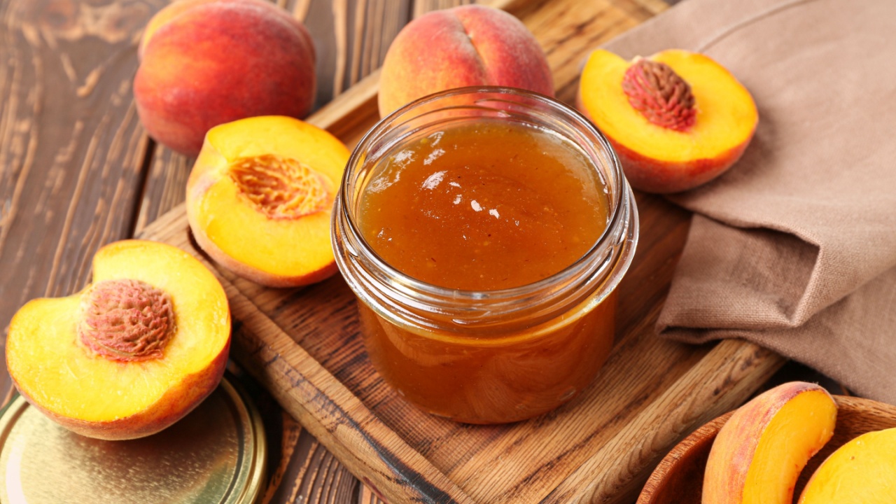 Cutting board with jar of sweet peach jam and fresh fruits on wooden background, closeup