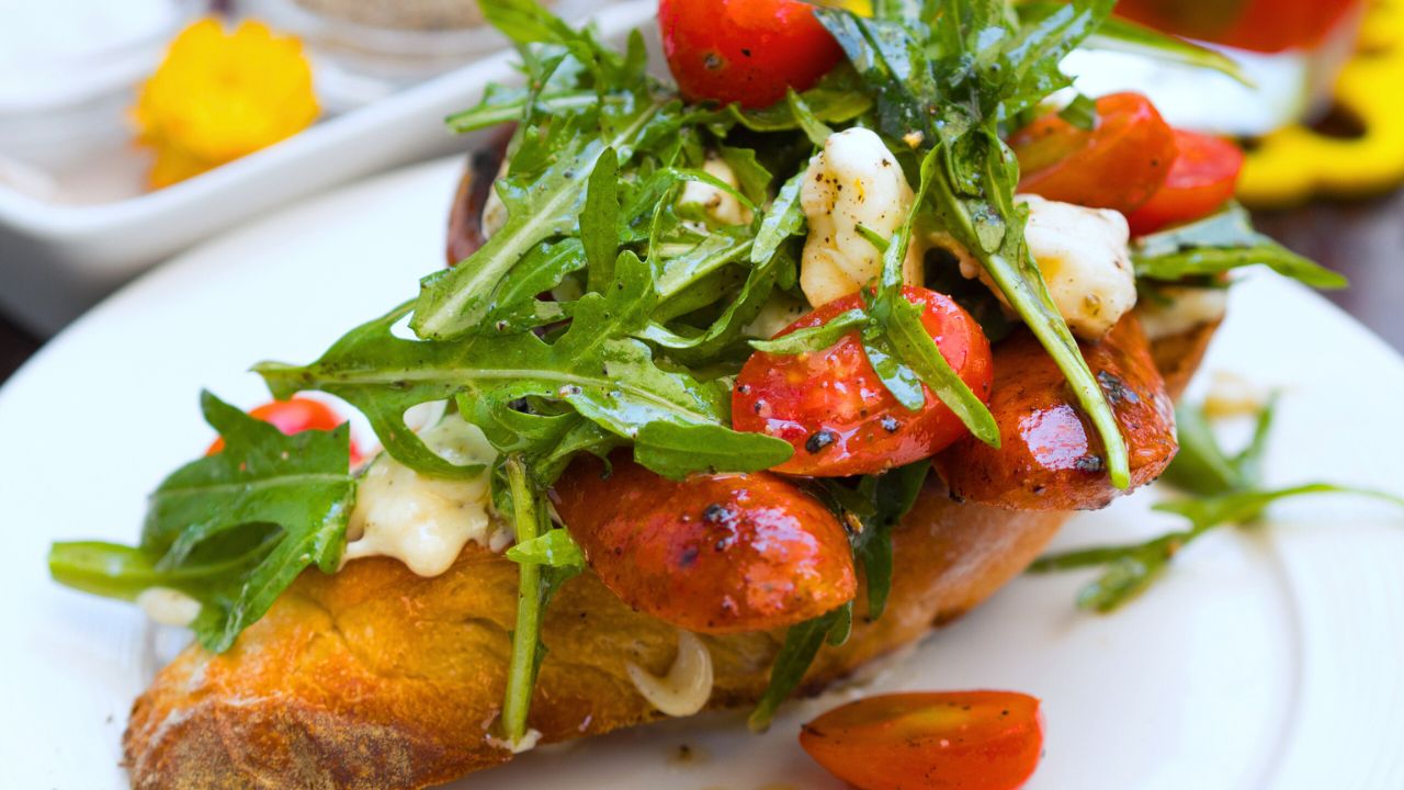 An open sandwich of toasted baguette with Gorgonzola, grilled chorizo, rocket salad, and tomatoes
