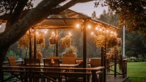 A cozy outdoor gazebo with hanging plants and string lights at dusk, surrounded by greenery, creating a warm and inviting atmosphere