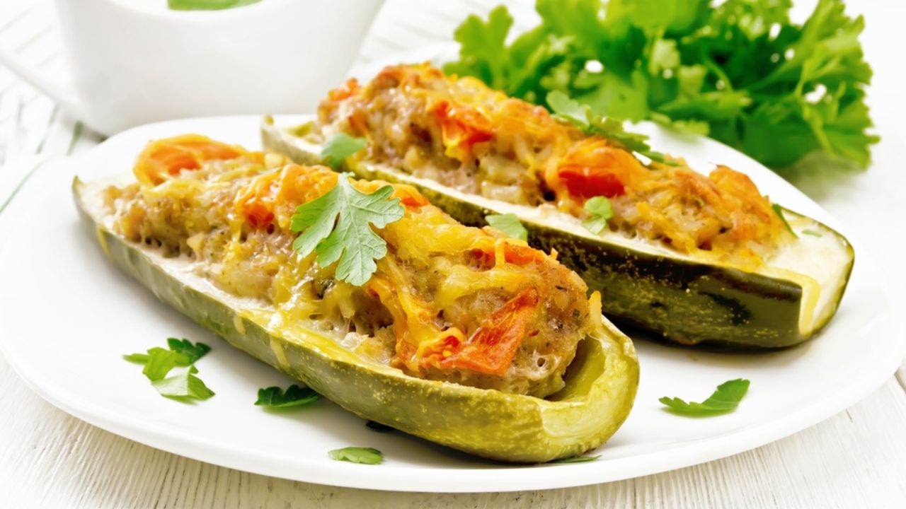 Cucumbers stuffed with minced meat, tomatoes, pepper with cheese and parsley in a plate, cream and greens sauce in a gravy boat, towel on white wooden board background