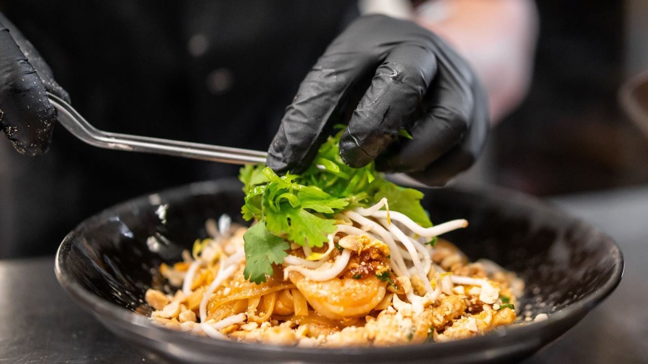 Chef plating Pad Thai in black bowl with shrimp, noodles, bean sprouts, peanuts, and cilantro garnish. Close-up of hands in gloves using tongs. Professional food presentation Chef plating Pad Thai in black bowl with shrimp, noodles, bean sprouts, peanuts, and cilantro garnish. Close-up of hands in gloves using tongs. Professional food presentation