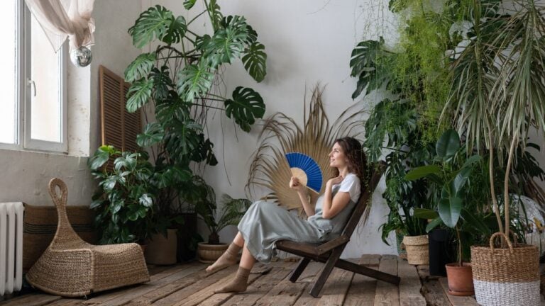 Indoor tropical oasis. Young relaxed dreamy woman using paper fan while escaping summer heat in beautiful and refreshing home garden full of large-leaved exotic plants, dreaming about vacation