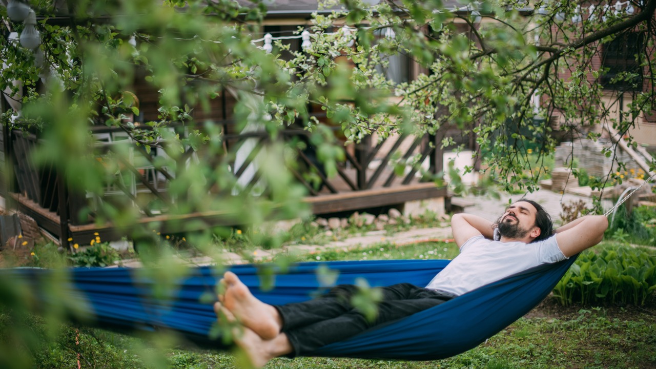 A man is resting in a hammock in a country house. A young guy lies in a hammock, dozing outdoors in the summer in the green of trees in the country