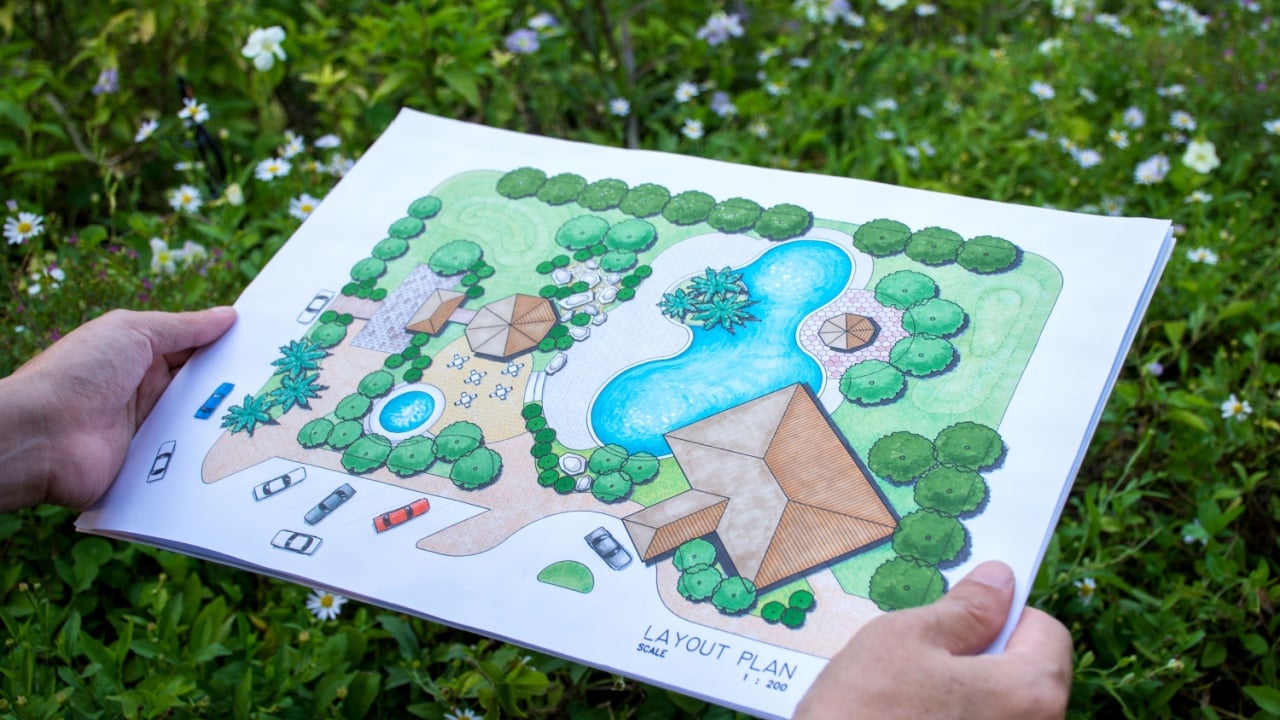 paper sheet of layout plan with hands and ีbush background, that shown design of clubhouse landscape or garden design drawing by hand with color marker pens and color pencil, selective focus