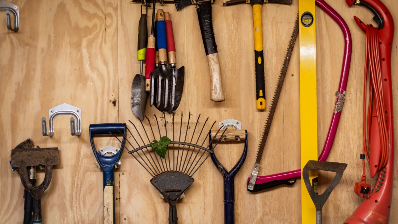 Assortment of DIY gardening tools and equipment hanging organised on wooden wall inside garden shed. Tools include rake, shovel, hammer, fork, trowel, spirit level measure, saw, axe, hatchet etc.
