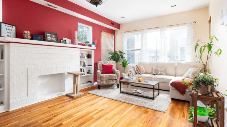 A bright living room with a red accent wall, white decorative fireplace, and natural hardwood floors