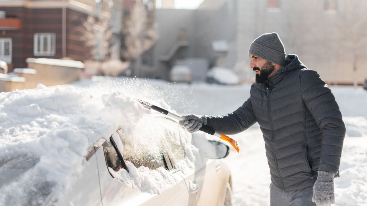 A young man cleans his car after a snowfall on a sunny, frosty day. Cleaning and clearing the car from snow on a winter day. Snowfall, and a severe snowstorm in winter.