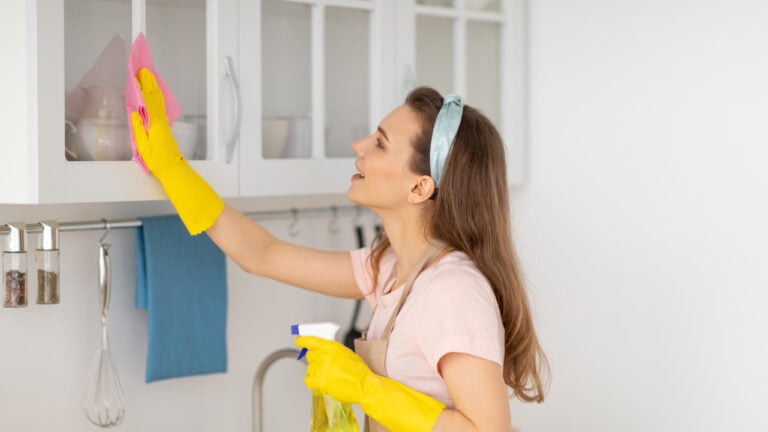 Lovely millennial housemaid wiping kitchen cabinet, performing house cleanup. Joyful young lady keeping her home tidy, doing domestic duties, using detergent and rag, side view