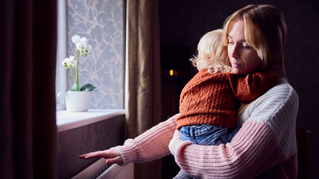 Mother With Son Trying To Keep Warm By Radiator At Home During Cost Of Living Energy Crisis