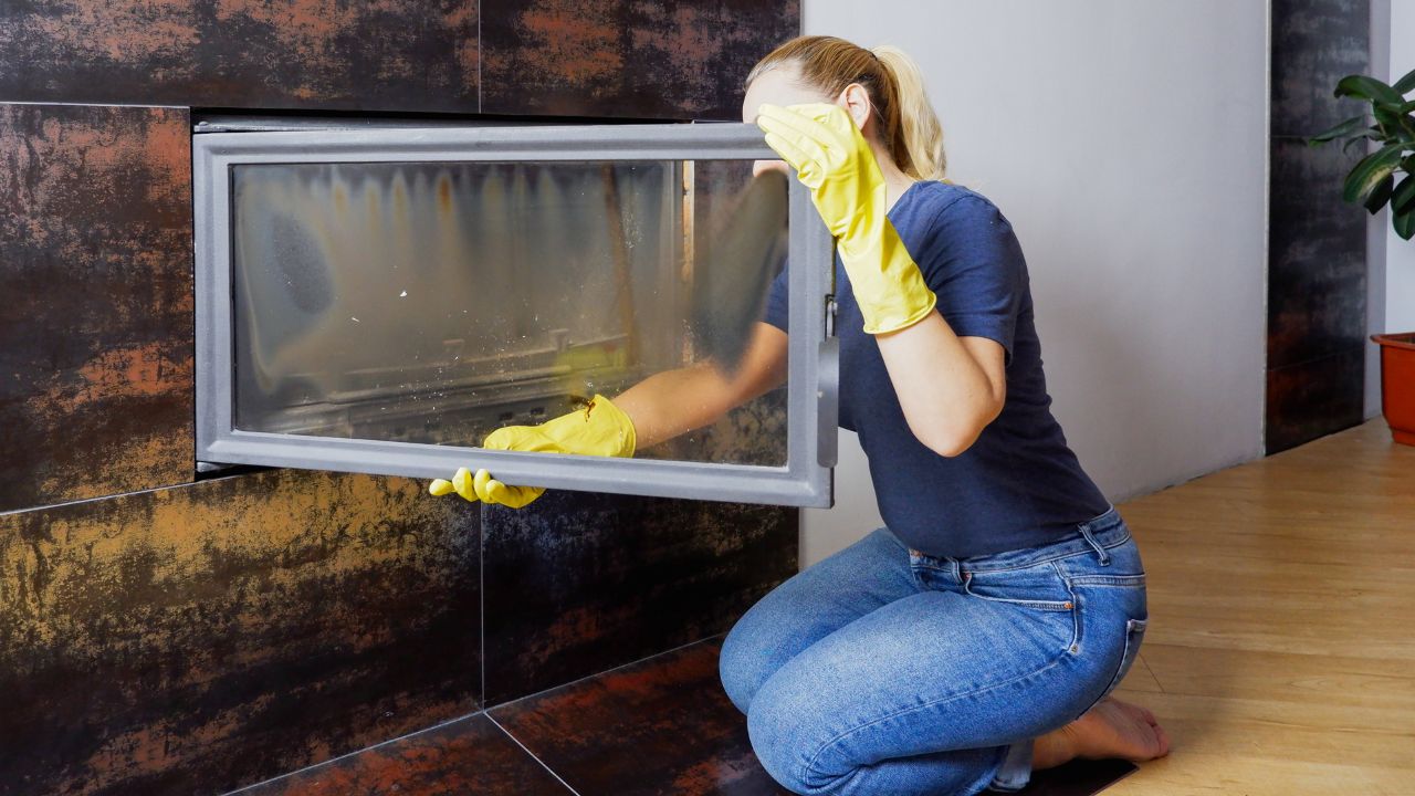 Close up of young woman in jeans, yellow gloves is cleaning the fireplace. Modern fireplace with glass.