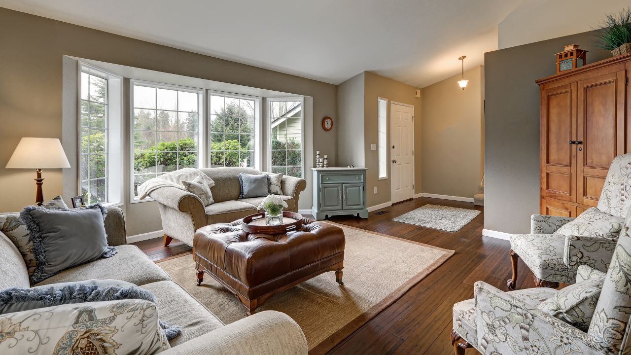 Cozy family room interior with taupe walls, sloped ceiling and wide plank wood floors. The room is furnished with claw foot chairs facing comfortable sofas across from brown leather tufted ottoman.