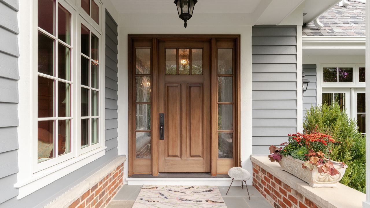 A front door and covered porch detail on a home with grey siding, white trim, red brick, a wooden front door.