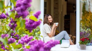 Smiling beautiful middle-aged woman with a cup of hot drink on the windowsill in the garden with blooming lilac