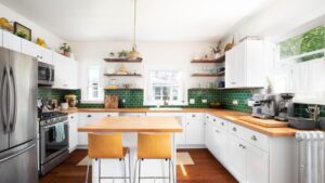A renovated kitchen with white cabinets, a natural wood countertop, chairs sitting at the island, a green subway tile backsplash, and a gold pendant light.