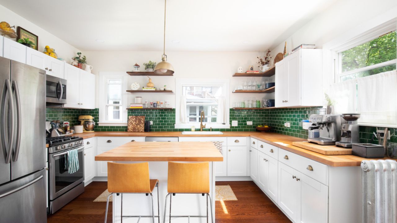 A renovated kitchen with white cabinets, a natural wood countertop, chairs sitting at the island, a green subway tile backsplash, and a gold pendant light.