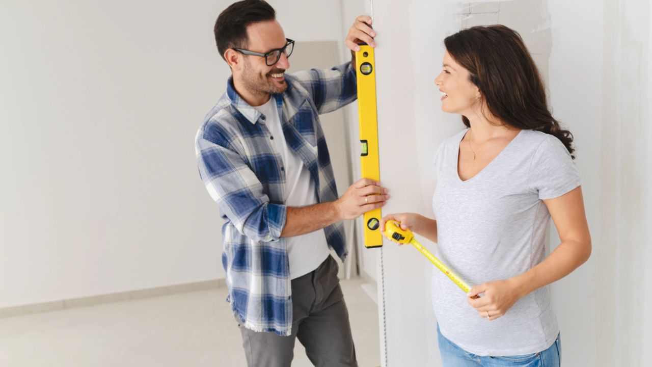 A young, beautiful, and cheerful couple, comprising a man and his pregnant wife, are measuring the walls, as part of their new home renovation project