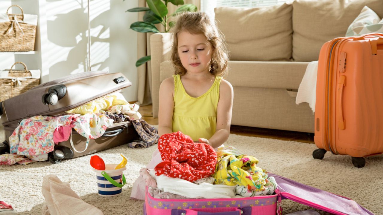 Happy little girl sitting in living room at home, carefully packing colorful summer clothes and toys into luggage suitcase for a new journey. Child Traveling