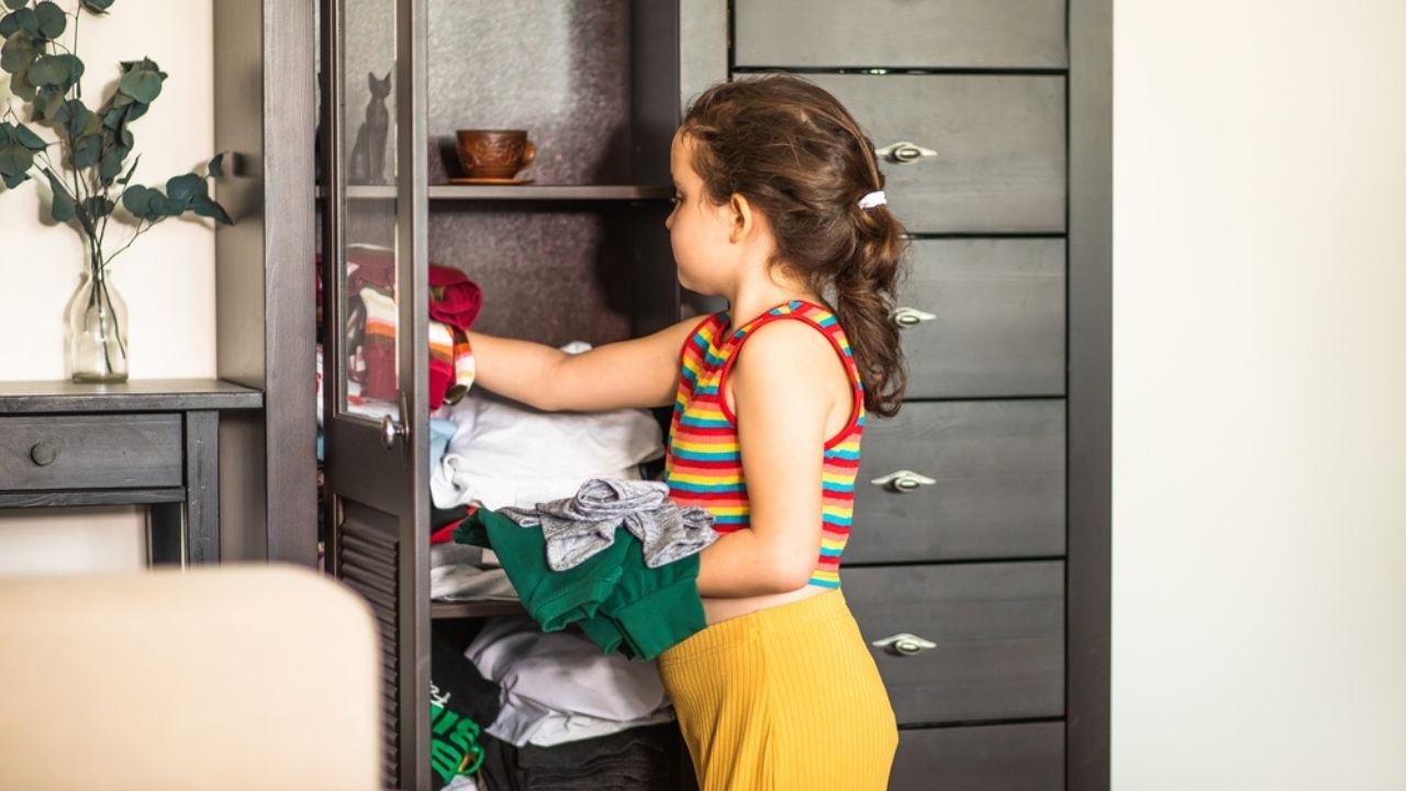 Kid holding stack pile of clothes near by wardrobe. Home chores housework concept.