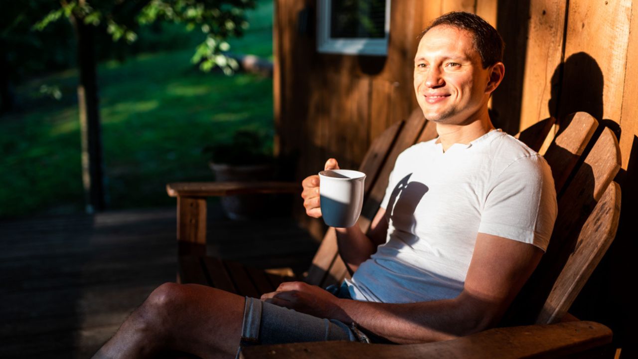 Man happy sitting relaxing on rocking chair lounge on porch of house in morning wooden cabin cottage drinking coffee or tea from cup mug