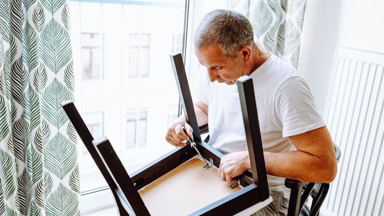 attractive middle-aged man, in white T-shirt, screwing screws in chair with screwdriver, repairing home furniture. Household chores, duties, furniture repair, broken woodwork