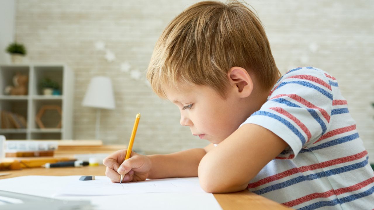 Side view portrait of diligent little boy writing or drawing carefully sitting at desk and doing homework, copy space