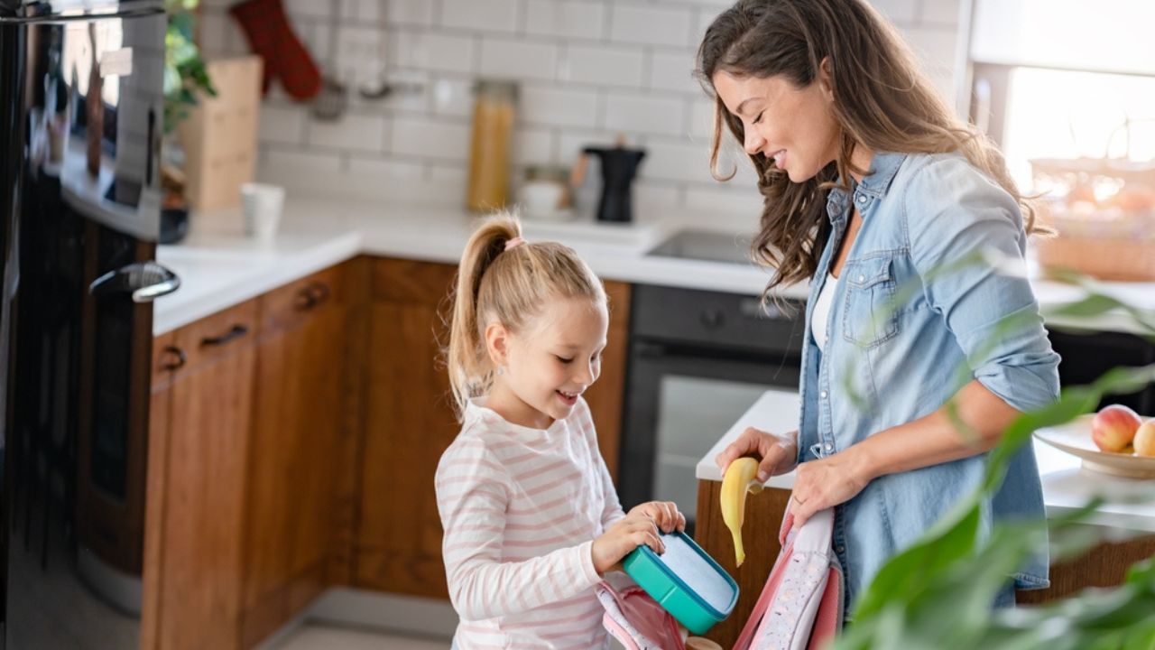 The cute girl is standing next to her caring young mother, who is preparing her to go to school giving her a fruit snack in a lunch box