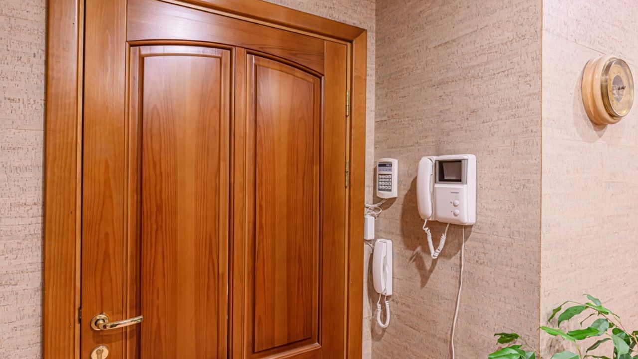 Warm, inviting entryway with a wooden door, intercom system, and textured walls. A round barometer and green plant add a touch of home.