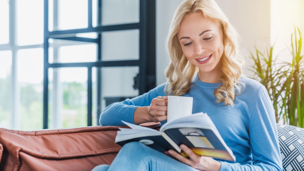 Young woman at home sitting on modern sofa relaxing in living room while reading book and drinking coffee