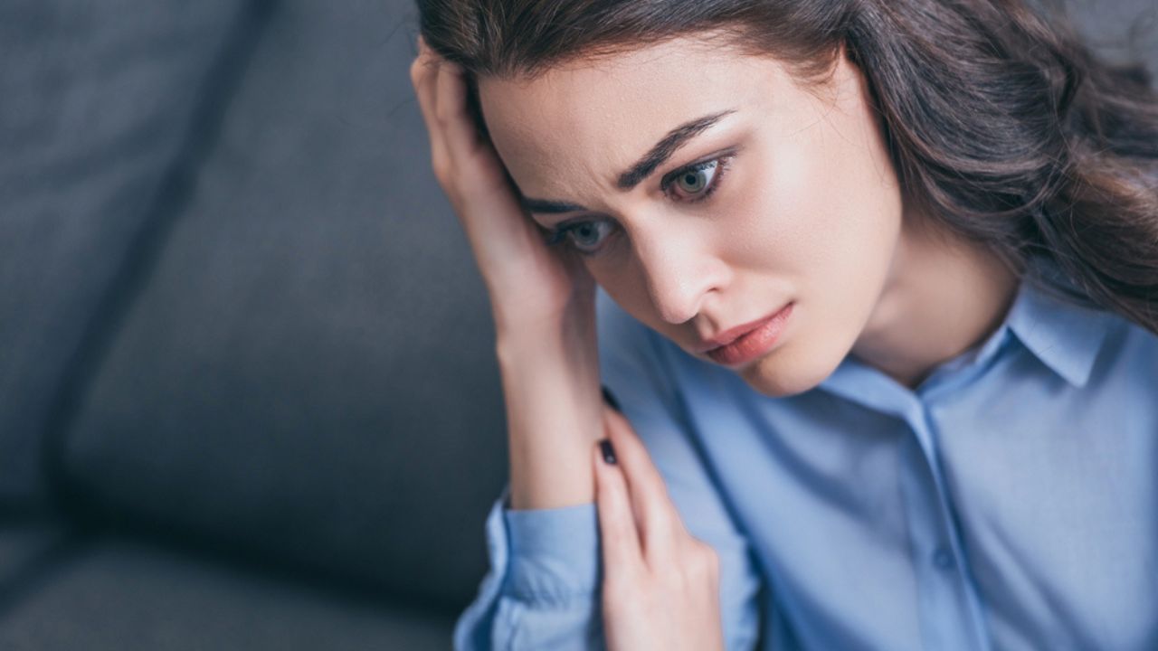 A contemplative young woman sits on a gray sofa, her expression reflecting deep thought or concern.