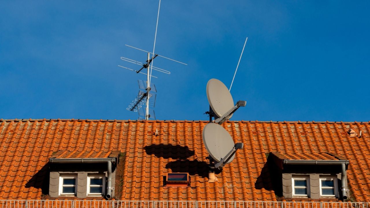 A tiled rooftop features two large satellite dishes alongside an antenna against a bright blue sky.