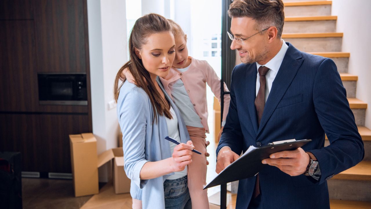 Attractive woman holding in arms daughter and signing document near handsome broker holding clipboard