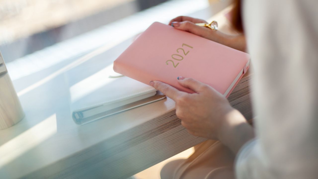 Female hands holding pink or coral coloured leather diary 2021 and pen while sitting near a window Concept of planning personal future goals and ideas for new year 2021. Copy space.