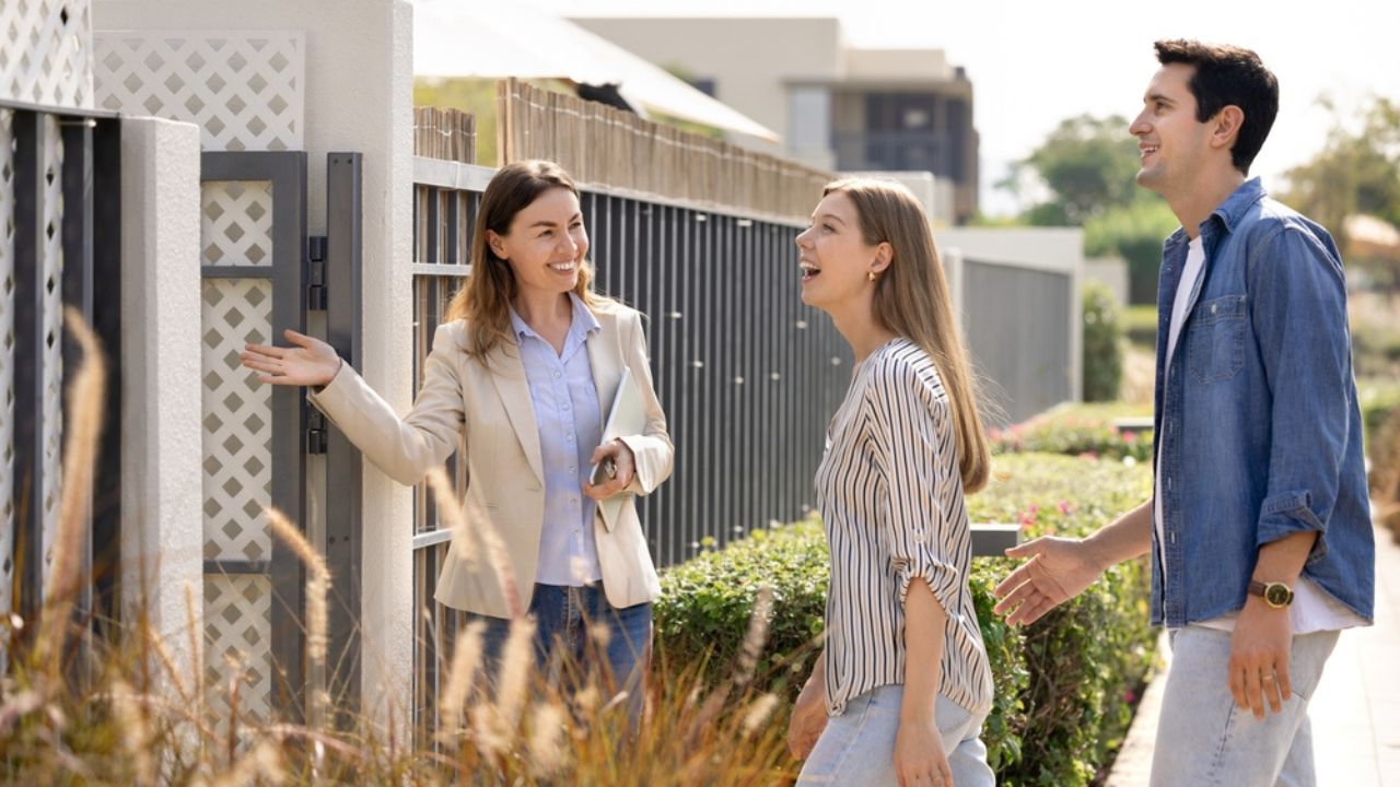 Friendly realtor agent welcoming young excited Hispanic married couple at entrance of modern residential property, showcasing new home for sale or rent.