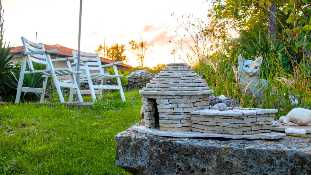 Home Garden design. Model of a stone house on a wall, behind a cat sculpture and two garden chairs in background