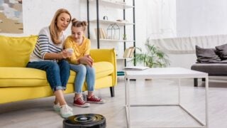 Mother and daughter looking at smartphone on sofa near coffee table with laptop and robotic vacuum cleaner on floor in living room