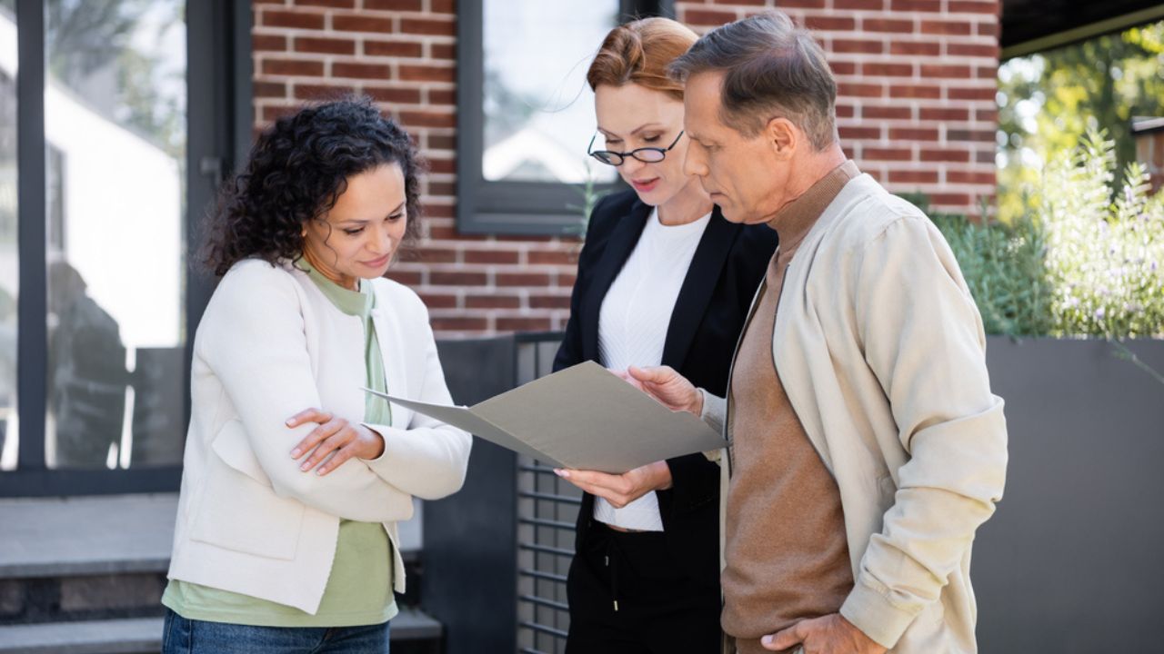 Multiethnic couple looking at folder near realtor and modern house outside