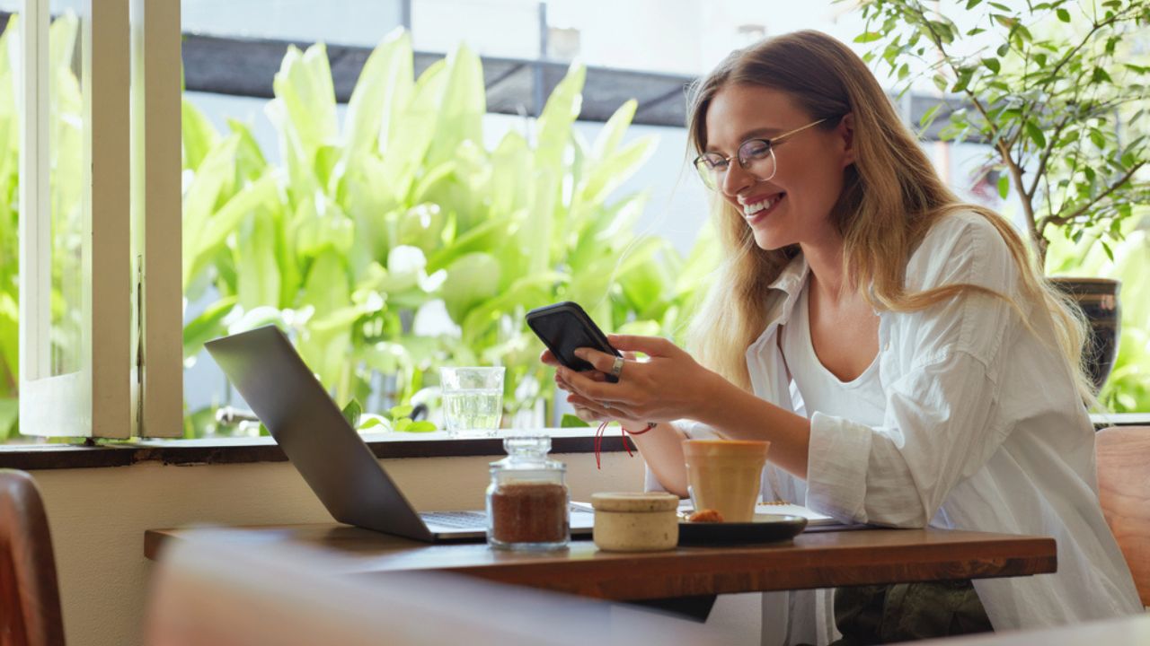 Online Job. Girl Works on Laptop and Using Smartphone at Cafe. Beautiful Business Woman in Casual Clothes and Glasses Portrait.