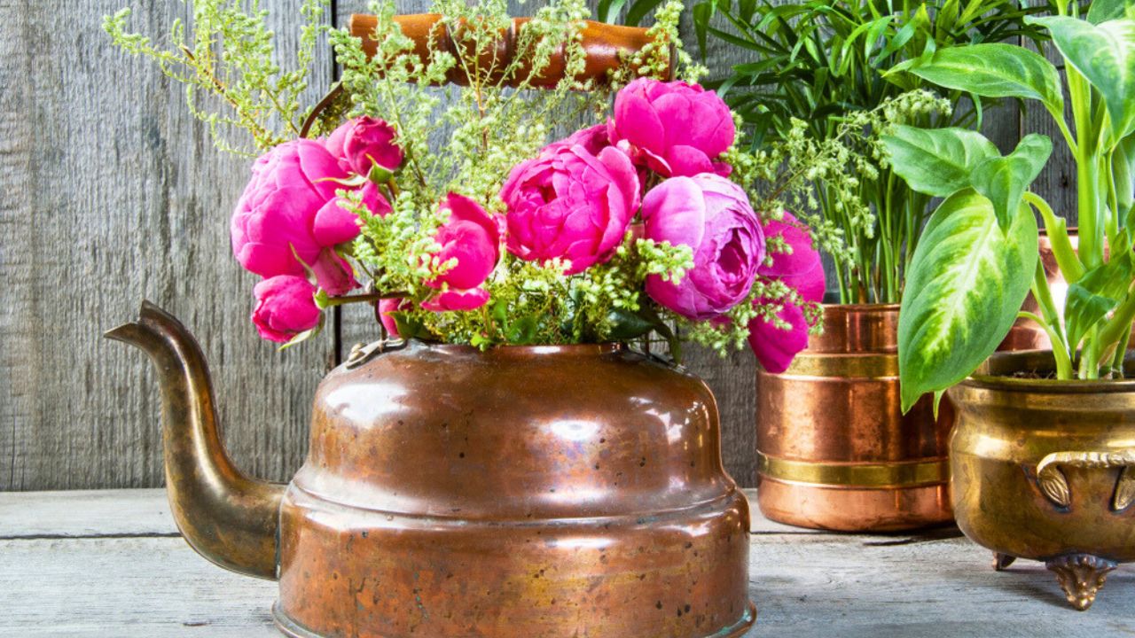 Pink roses in old copper brass kettle with wooden handle and green plants in vintage flower pot cpose up on wooden background. Copy space for text.