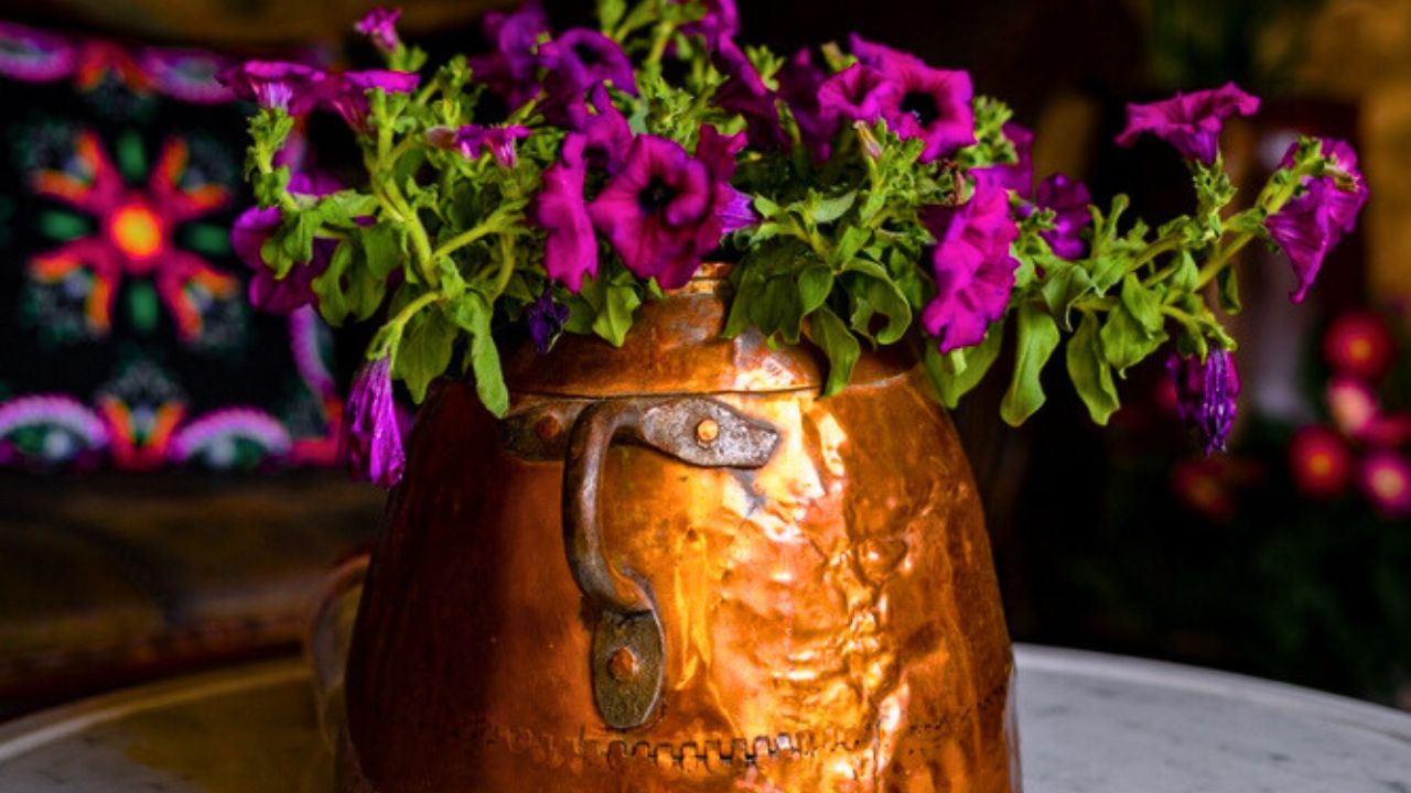 Rustic copper pot with vibrant purple petunias placed on a marble table in a cozy interior with stone wall and traditional textiles, evoking a charming and vintage atmosphere.