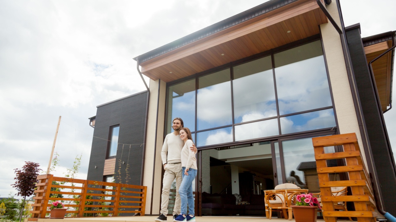 Happy young couple embracing standing outside big house on front porch, smiling owners enjoying luxury real estate wood and glass exterior design, buying home, mortgage loan, vacation rentals concept