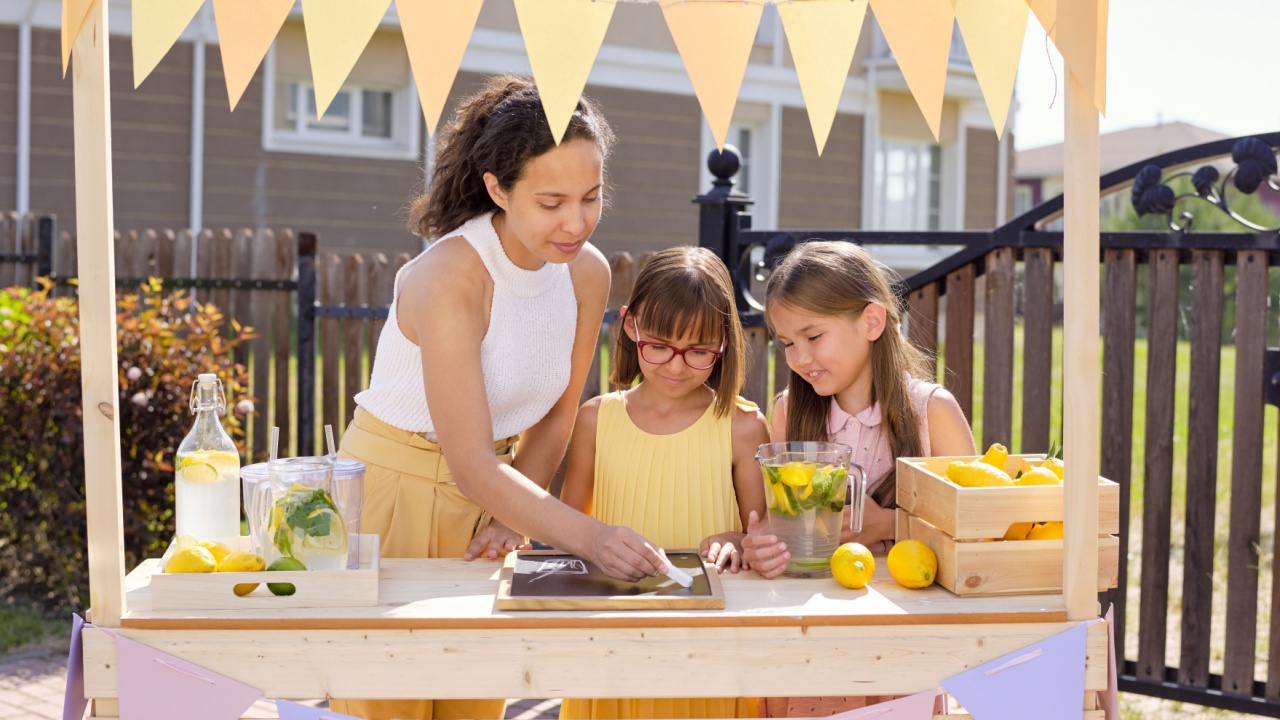 Young brunette woman drawing glass of drink on noticeboard with chalk while her daughters standing near by and selling lemonade by stall