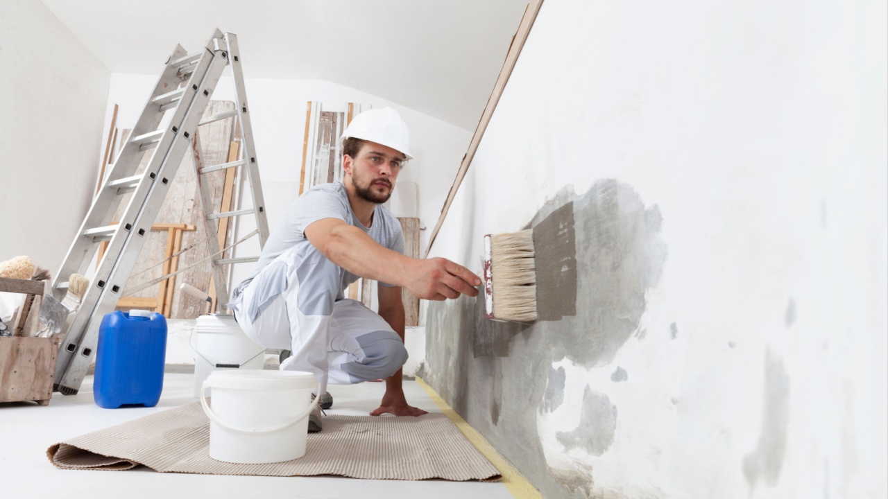 construction worker painter man with protective helmet, brush in hand and buckets of products to restore and paint the wall, indoor the building site of a house