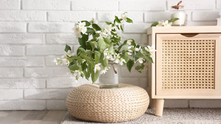Bouquet of beautiful jasmine flowers in glass vase near white brick wall indoors