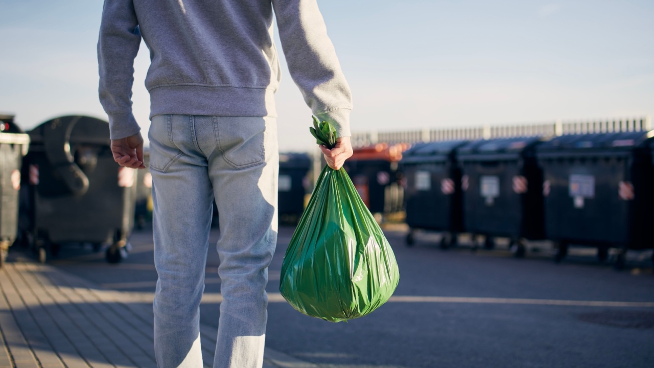 Man walking with rubbish. Rear view of person carrying plastic bag against garbage cans on street.