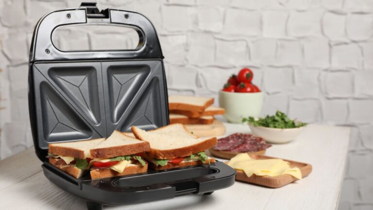 Modern grill maker with tasty sandwiches and ingredients on white wooden table