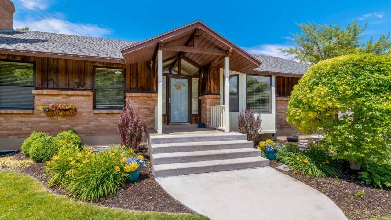 Facade of home with paved walkway leading to the portico and white front door. Front yard with lush foliage and flowers, stone wall, sidelight, and outdoor stairs can also be seen at the exterior.
