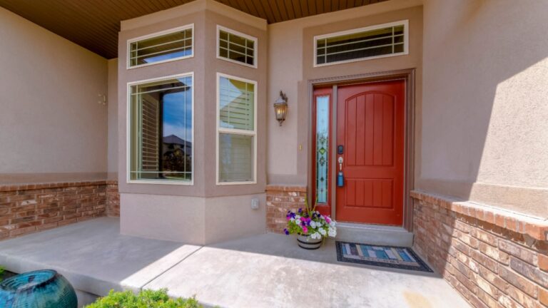 Entrance of a house with red front door with lockbox, sidelight, and transom window. Home exterior with bay windows near the flowers on the right at the front of the sidelight with ornate glass.