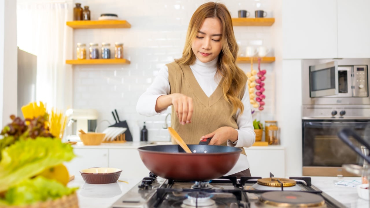 Asian woman cooking healthy food fried pasta in cooking pan on modern kitchen island stove at home. Attractive girl preparing food for dinner party celebration meeting with friend on holiday vacation.
